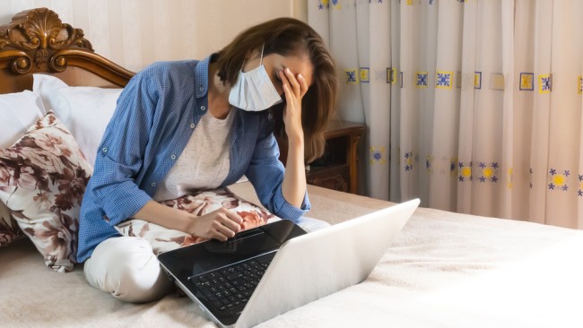 Young woman in medical mask after online work at home tired sitting on bed — Stock Image
